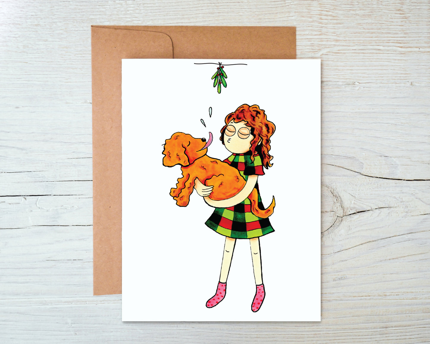 A greeting card featuring a cartoon girl in a Christmas dress holding a golden doodle puppy as it licks her face underneath a mistletoe.