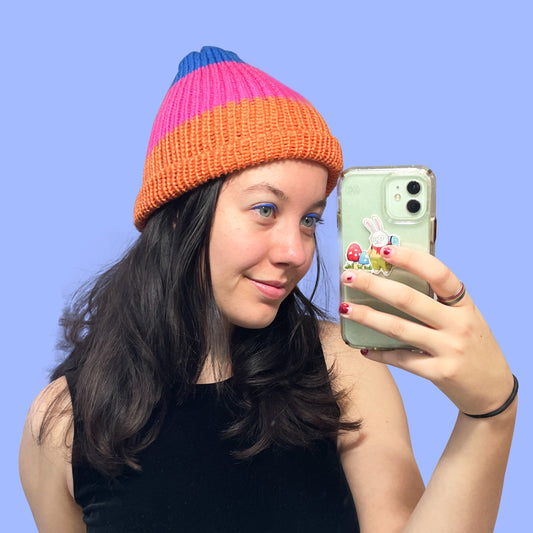 A white woman with dark brown hair wearing the orange-pink-blue color-block beanie and smiling in the mirror