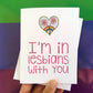 A hand holding a greeting card and the front reads, "I'm in lesbians with you" with an illustration of a intertwined pink flowers inside of a heart.