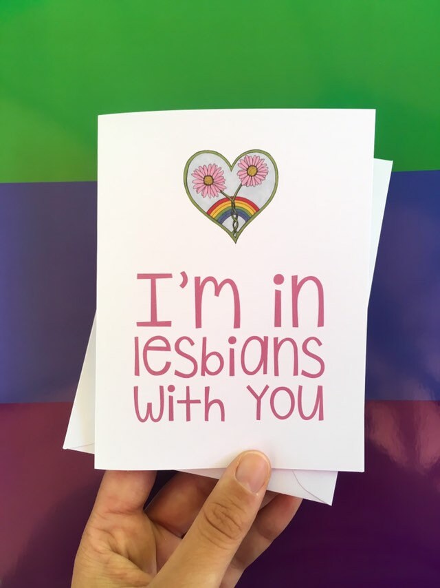 A hand holding a greeting card and the front reads, "I'm in lesbians with you" with an illustration of a intertwined pink flowers inside of a heart.