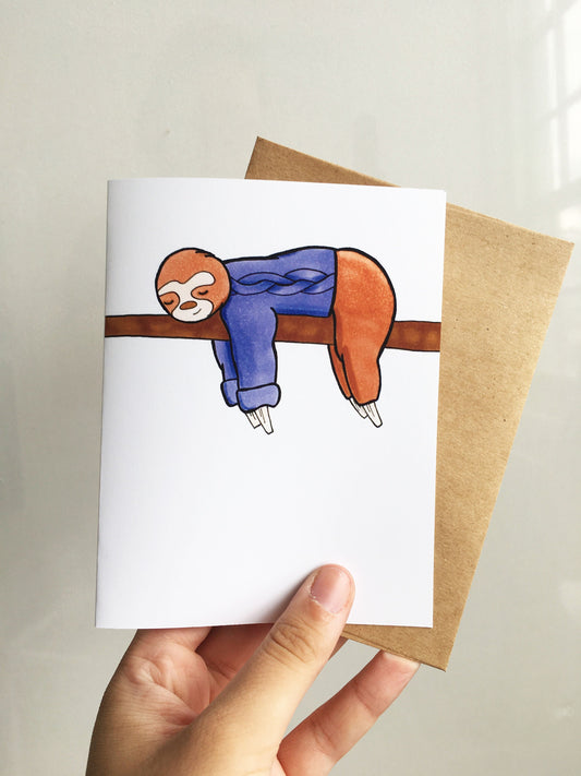 A hand holding a greeting card featuring an image of a cartoon sloth sleeping on a tree branch while wearing a purple knit sweater.