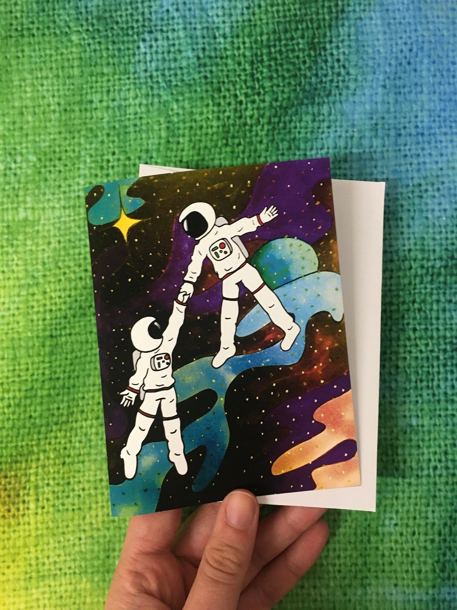 A hand holding a greeting card featuring an illustration of two astronauts floating in space while holding hands. The inside of the card reads, "you take me to outerspace."