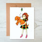 A greeting card featuring a cartoon girl in a Christmas dress holding a golden doodle puppy as it licks her face underneath a mistletoe.